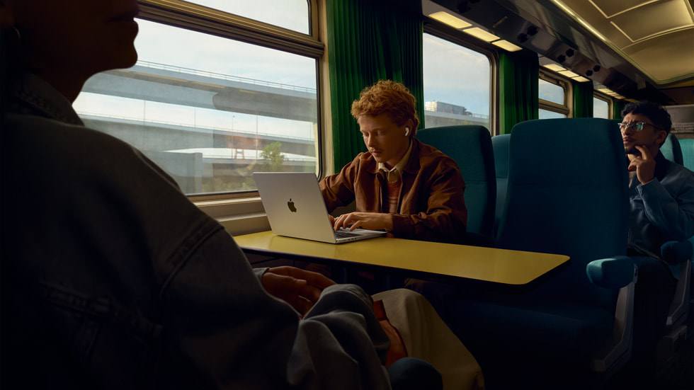 A traveler wearing AirPods Pro 3 while working on a MacBook aboard a train
